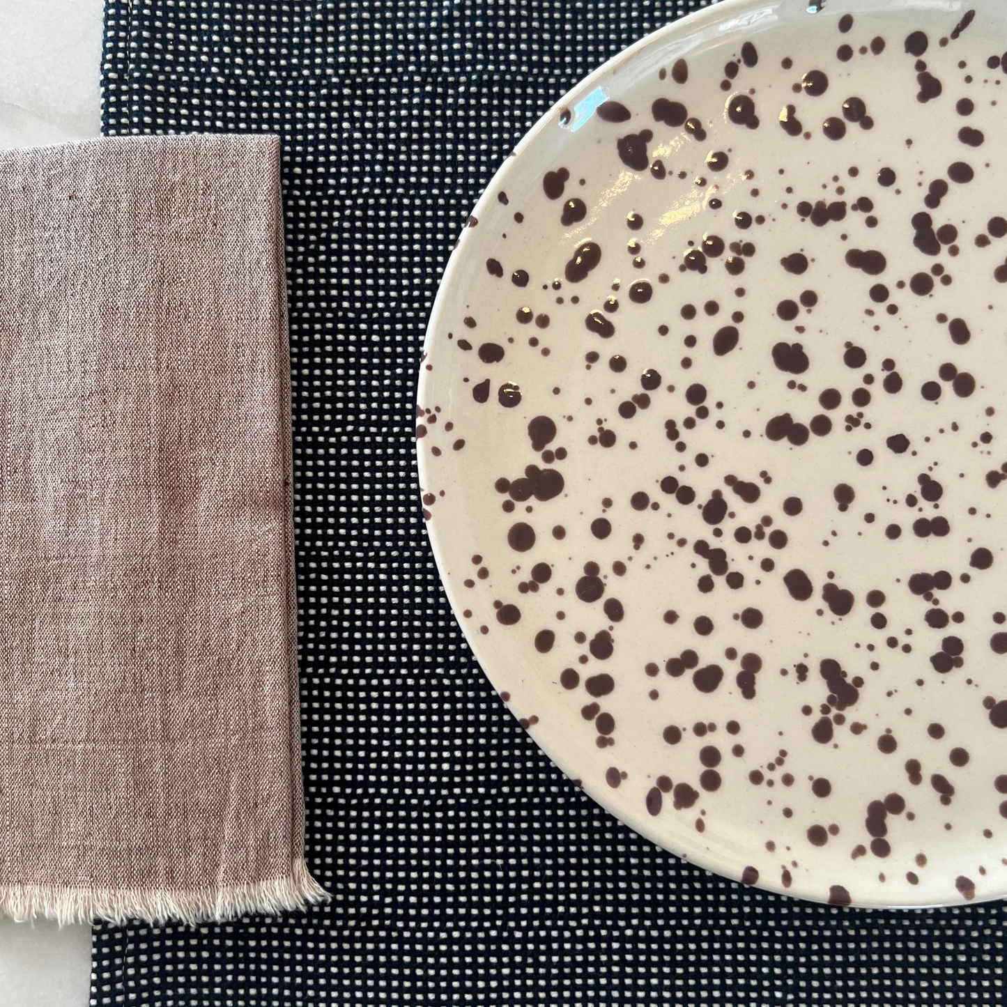Speckled ceramic plate on a navy placemat with a brown napkin on a marble surface