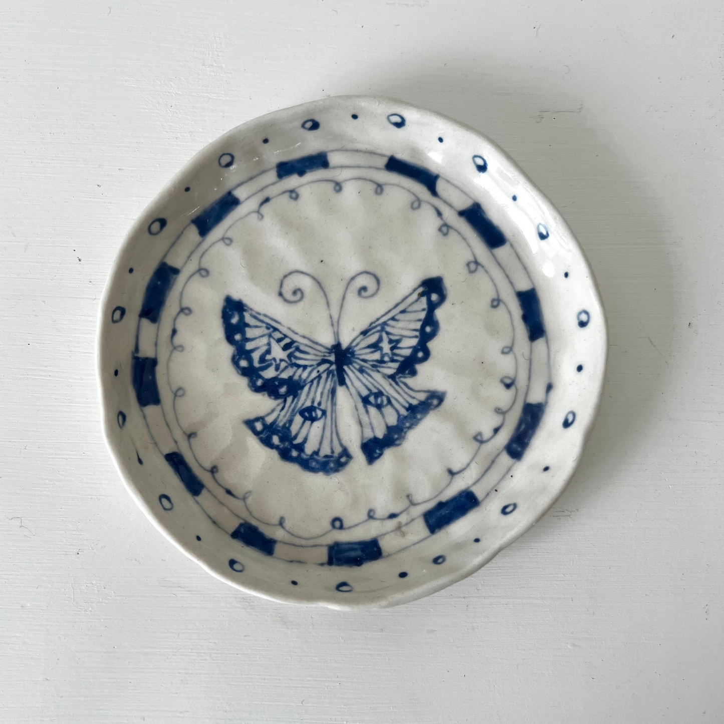A small round white ceramic dish with a blue butterfly design in the center and blue dotted border patterns, photographed from above on a white surface.