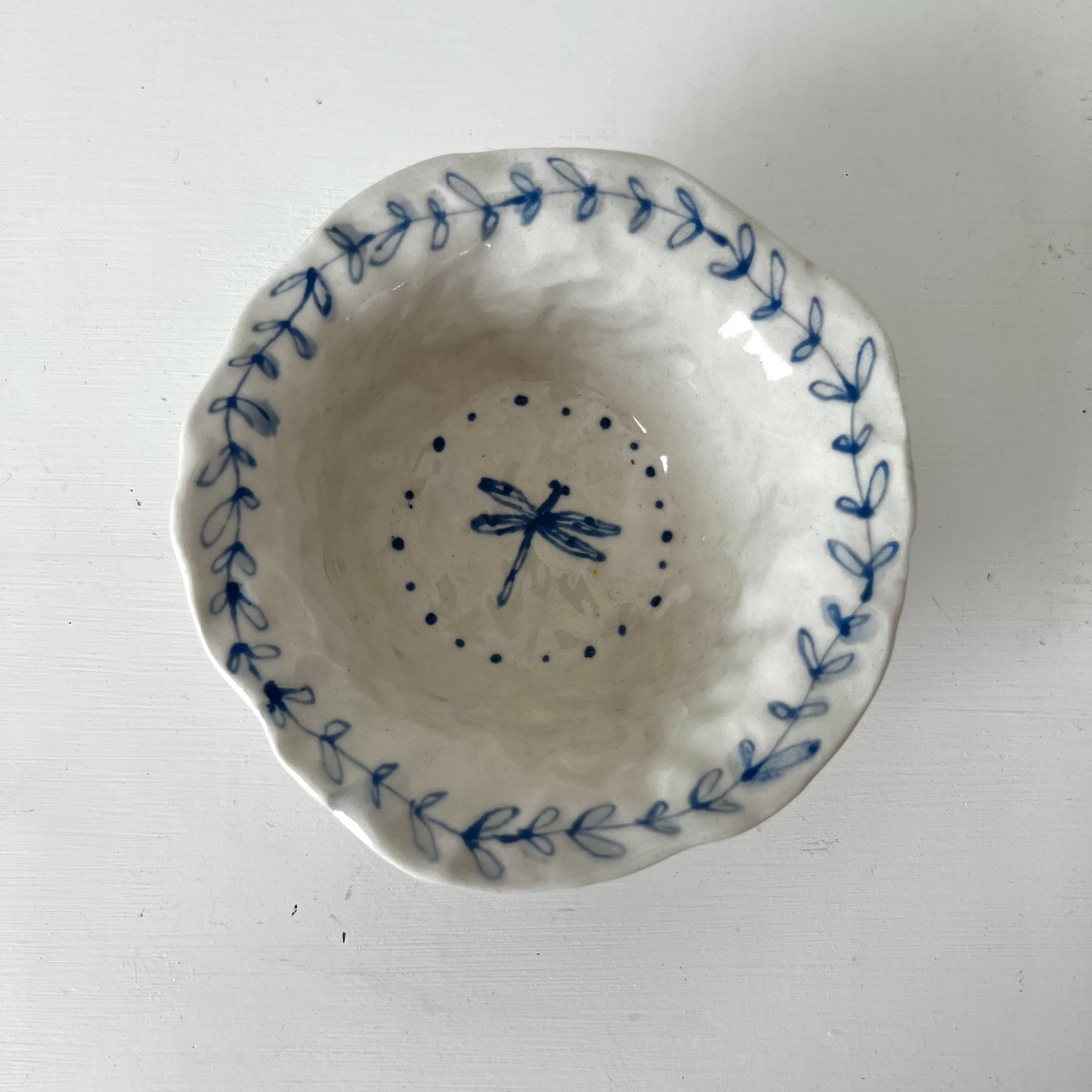 A small handmade ceramic bowl with a scalloped rim, featuring blue hand-painted leaf patterns around the edge and a blue dragonfly design in the center, shown from a top-down view on a white background.