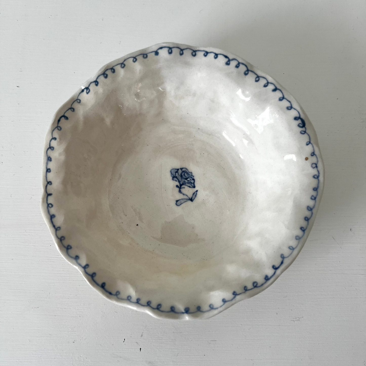 A white ceramic bowl with a scalloped rim decorated with a blue dotted pattern and a small blue floral motif in the center, shown from a top-down view on a white surface.