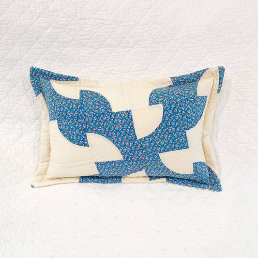 A square quilted throw pillow featuring a blue and cream patchwork design with a small floral print, displayed on a white quilted blanket.
