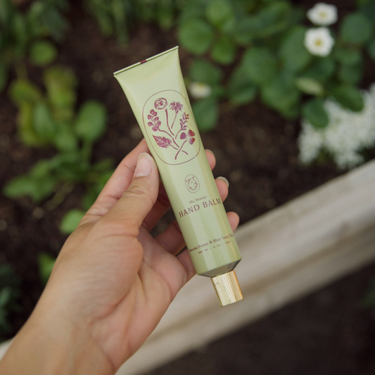 Hand holding a green tube of hand balm with floral design against a garden background