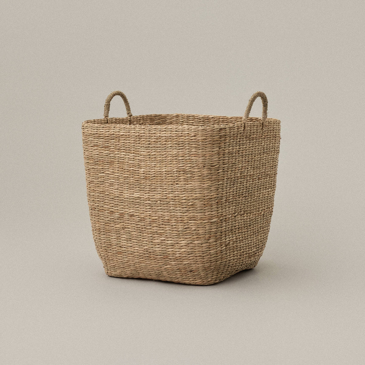 A natural woven seagrass storage basket with two loop handles, shown on a neutral warm gray studio background.