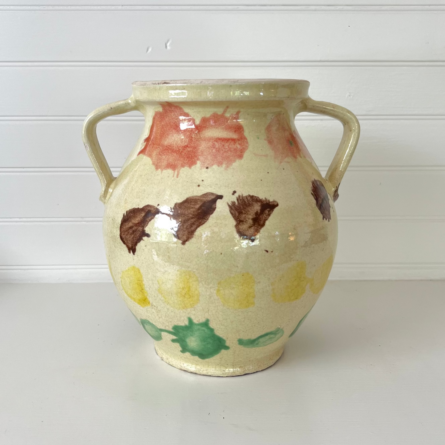 A handmade ceramic vase with two side handles, featuring a cream-colored glaze decorated with hand-painted abstract shapes in brown, red, yellow, and green, set against a white paneled background.