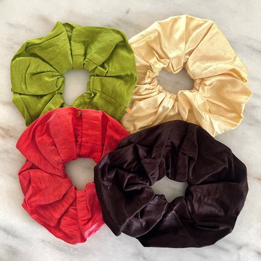 Four fabric hair scrunchies in olive green, cream, red, and dark brown velvet, arranged in a square on a white marble surface.