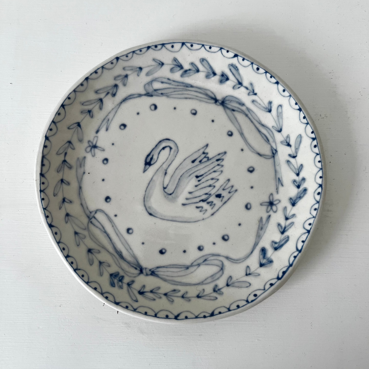 A round white ceramic plate with a blue hand-painted illustration of a swan, surrounded by a leafy wreath and dotted border, photographed from directly above on a white surface.