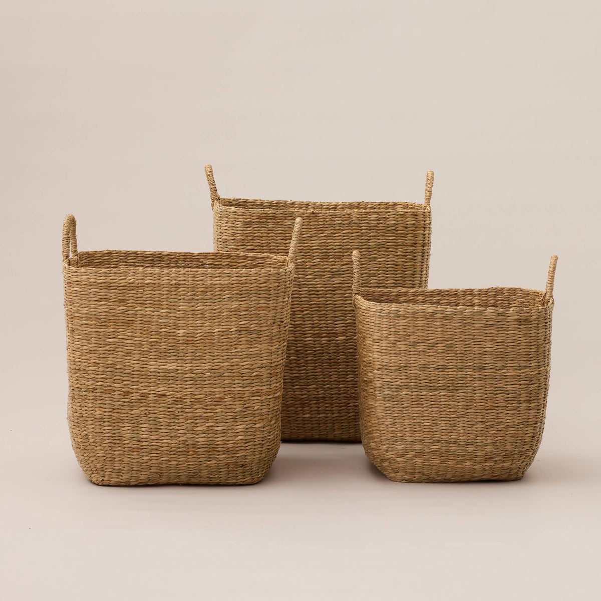 Three woven natural fiber storage baskets in different heights, arranged on a plain beige studio background.