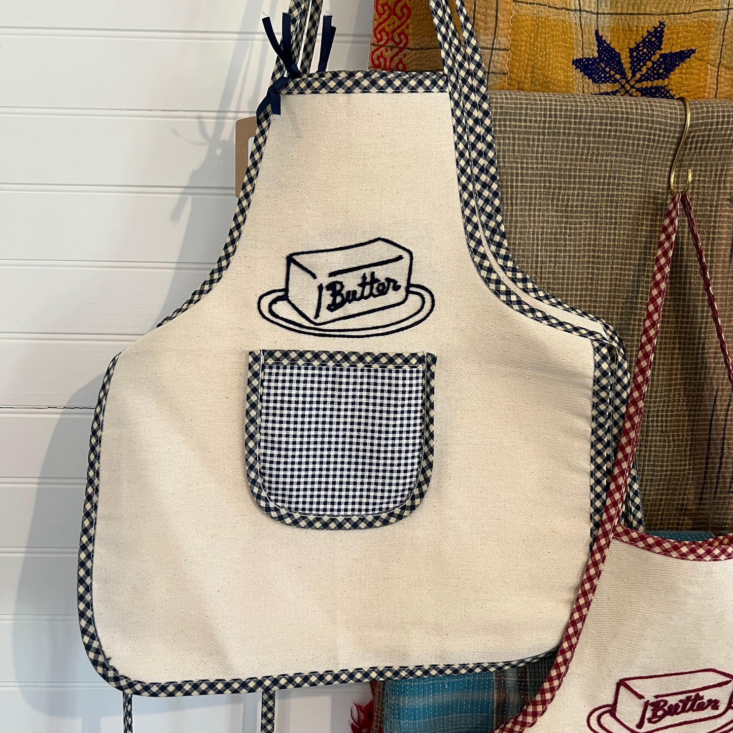 Beige apron with black checkered trim and 'Butter' design hanging on a hook.
