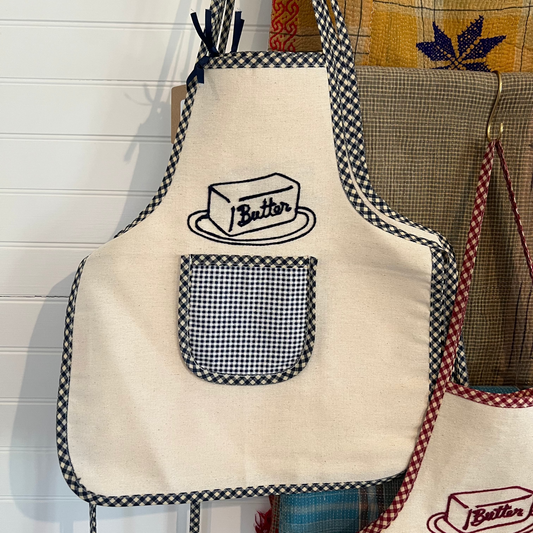 Beige apron with black checkered trim and 'Butter' design hanging on a hook.