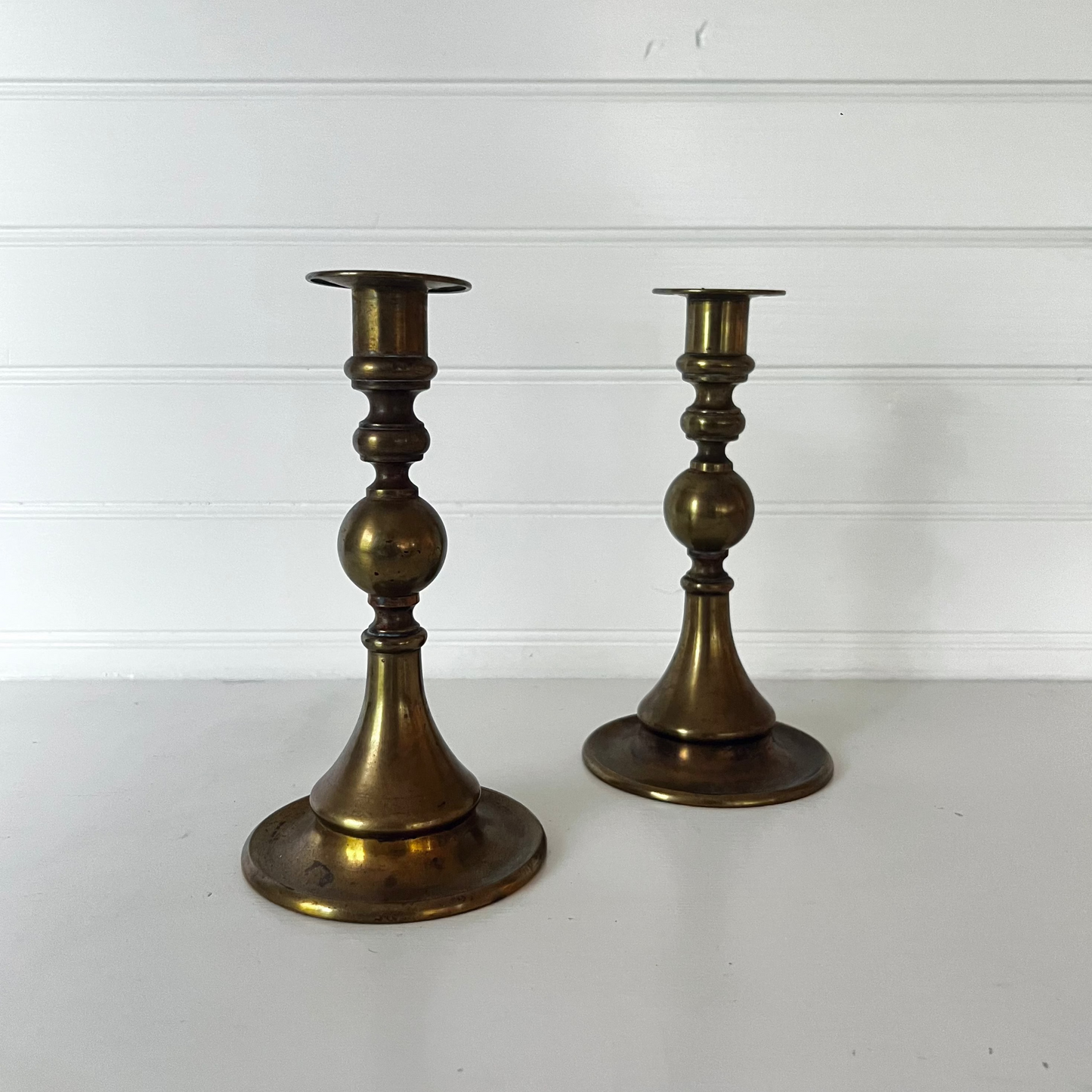 Two brass candlesticks on a white surface with a white wooden background
