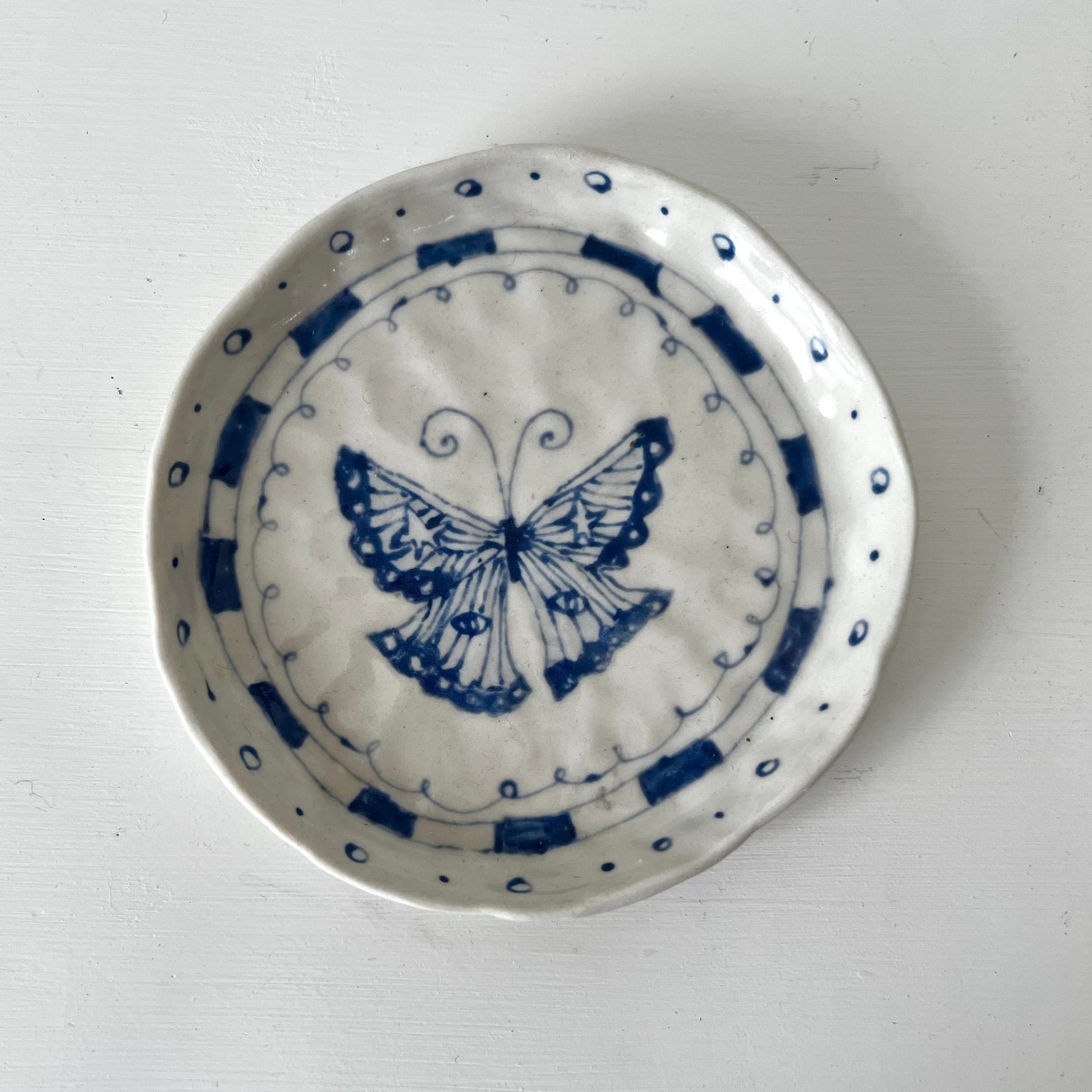 A small round white ceramic dish with a blue butterfly design in the center and blue dotted border patterns, photographed from above on a white surface.