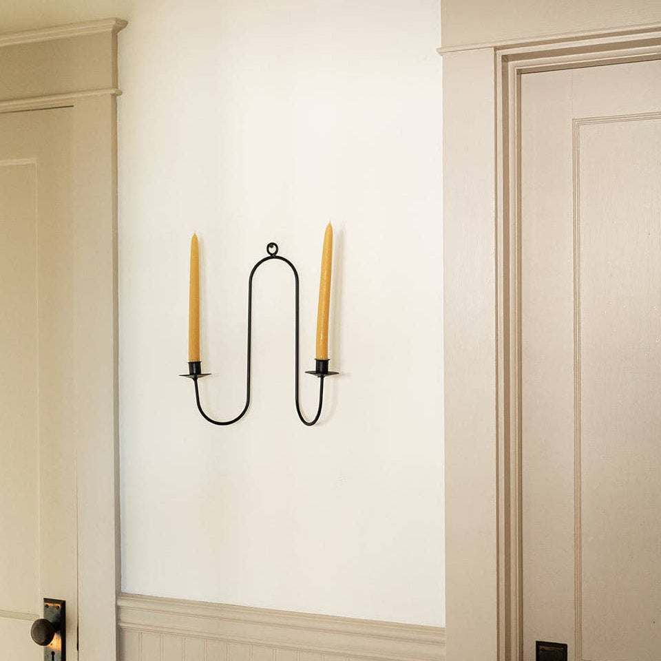A black double candle sconce mounted on a white wall between two doors, holding two yellow taper candles.