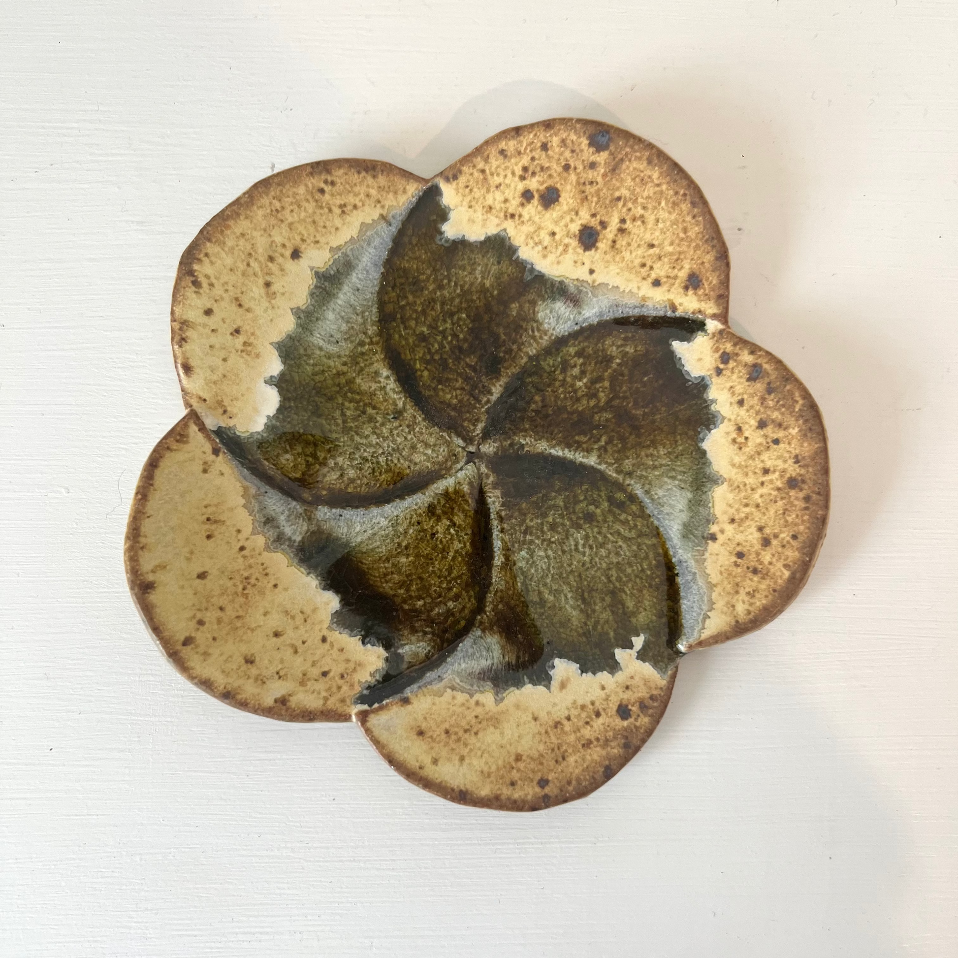 A handmade ceramic flower-shaped dish with a textured, speckled brown and beige glaze, photographed from directly above on a white surface.