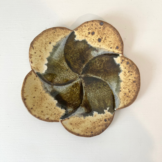 A handmade ceramic flower-shaped dish with a textured, speckled brown and beige glaze, photographed from directly above on a white surface.